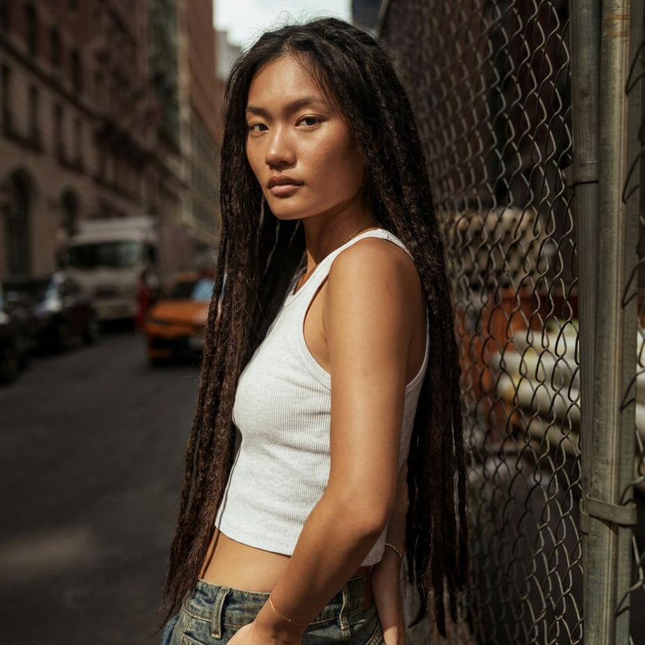 Community model wearing long dark straight loc extensions, a white ribbed tank top and denim jeans in a city street setting.
